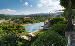 Scenic view of a luxury villa with an infinity pool overlooking rolling green hills and a blue sky, featuring sun loungers and landscaped gardens.