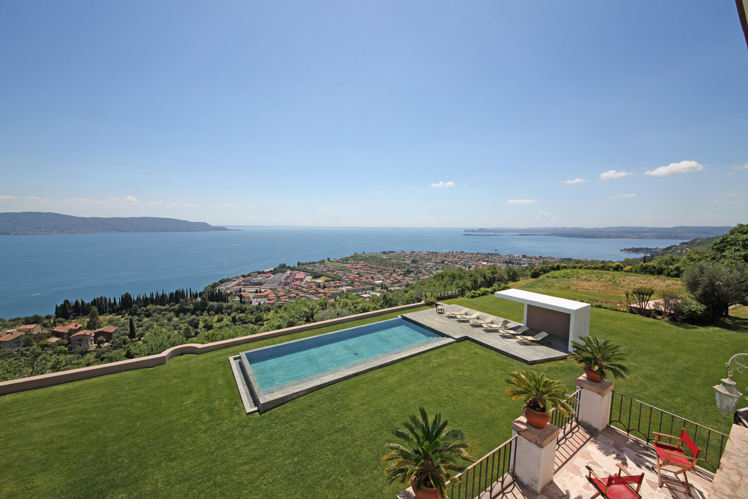 Scenic view of a modern villa with a swimming pool overlooking a coastal landscape, featuring lush greenery and a clear blue sky. | Only on SopranoVillas