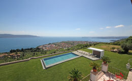 Scenic view of a modern villa with a swimming pool overlooking a coastal landscape, featuring lush greenery and a clear blue sky.