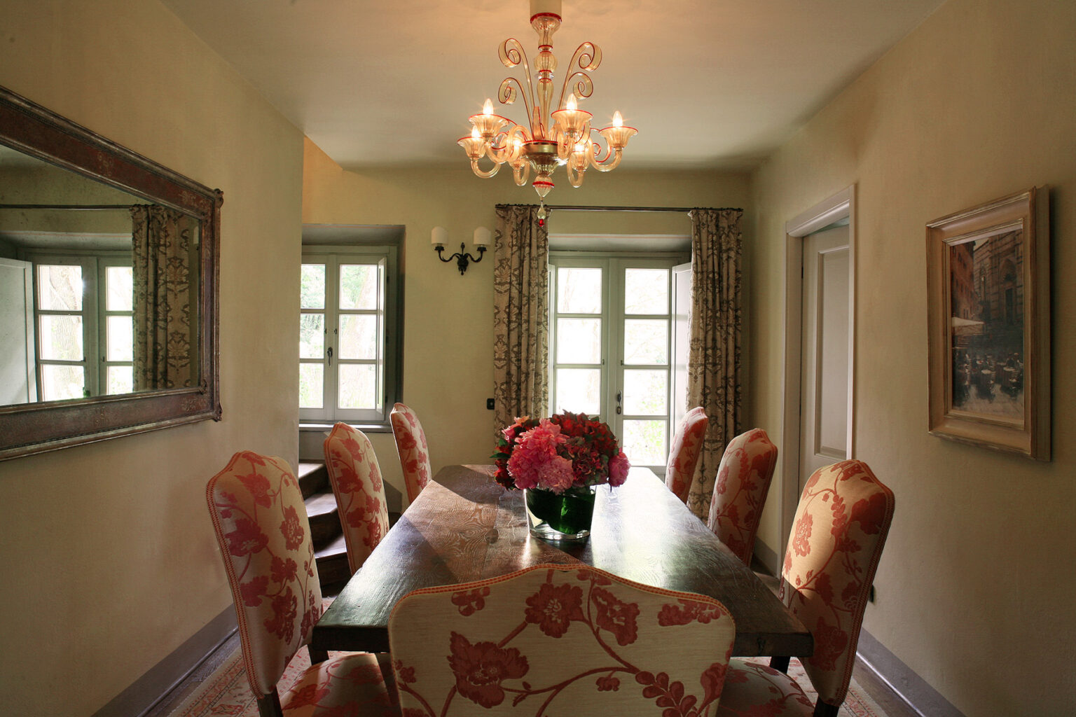Elegant dining room featuring a large wooden table surrounded by floral upholstered chairs, a decorative chandelier, and a mirror reflecting natural light from the windows. Fresh flowers in a vase add a touch of color to the inviting space. | Only on SopranoVillas