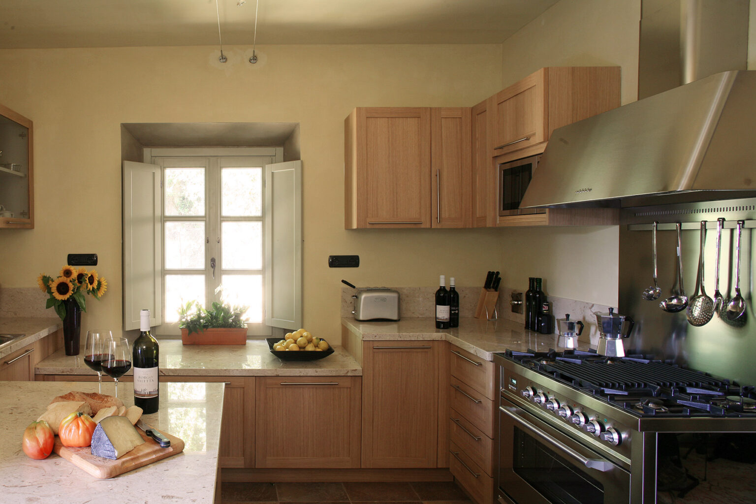 Modern kitchen with wooden cabinets, stainless steel appliances, and a window providing natural light. The countertop features fresh fruits, a bottle of wine, and glasses, creating a warm and inviting atmosphere. | Only on SopranoVillas