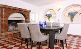Elegant dining room featuring a dark wooden table surrounded by plush gray chairs, with a cozy brick fireplace in the background. Natural light streams through arched windows adorned with blue hydrangea arrangements, enhancing the inviting atmosphere.