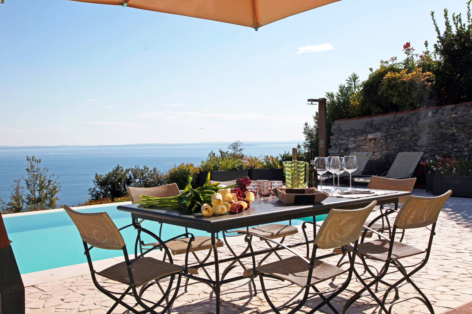 Elegant outdoor dining setup by a pool with a stunning ocean view, featuring a stylish table with fresh fruits, glasses, and a sun umbrella for shade. Ideal for alfresco dining and relaxation. | Only on SopranoVillas