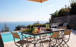 Elegant outdoor dining setup by a pool with a stunning ocean view, featuring a stylish table with fresh fruits, glasses, and a sun umbrella for shade. Ideal for alfresco dining and relaxation.