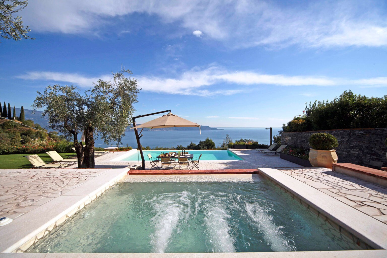 Luxury villa with a swimming pool overlooking a serene lake, surrounded by lush greenery and blue skies. The image showcases a relaxing outdoor space with sun loungers and a shaded dining area. | Only on SopranoVillas