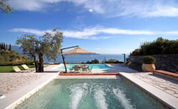 Luxury villa with a swimming pool overlooking a serene lake, surrounded by lush greenery and blue skies. The image showcases a relaxing outdoor space with sun loungers and a shaded dining area.