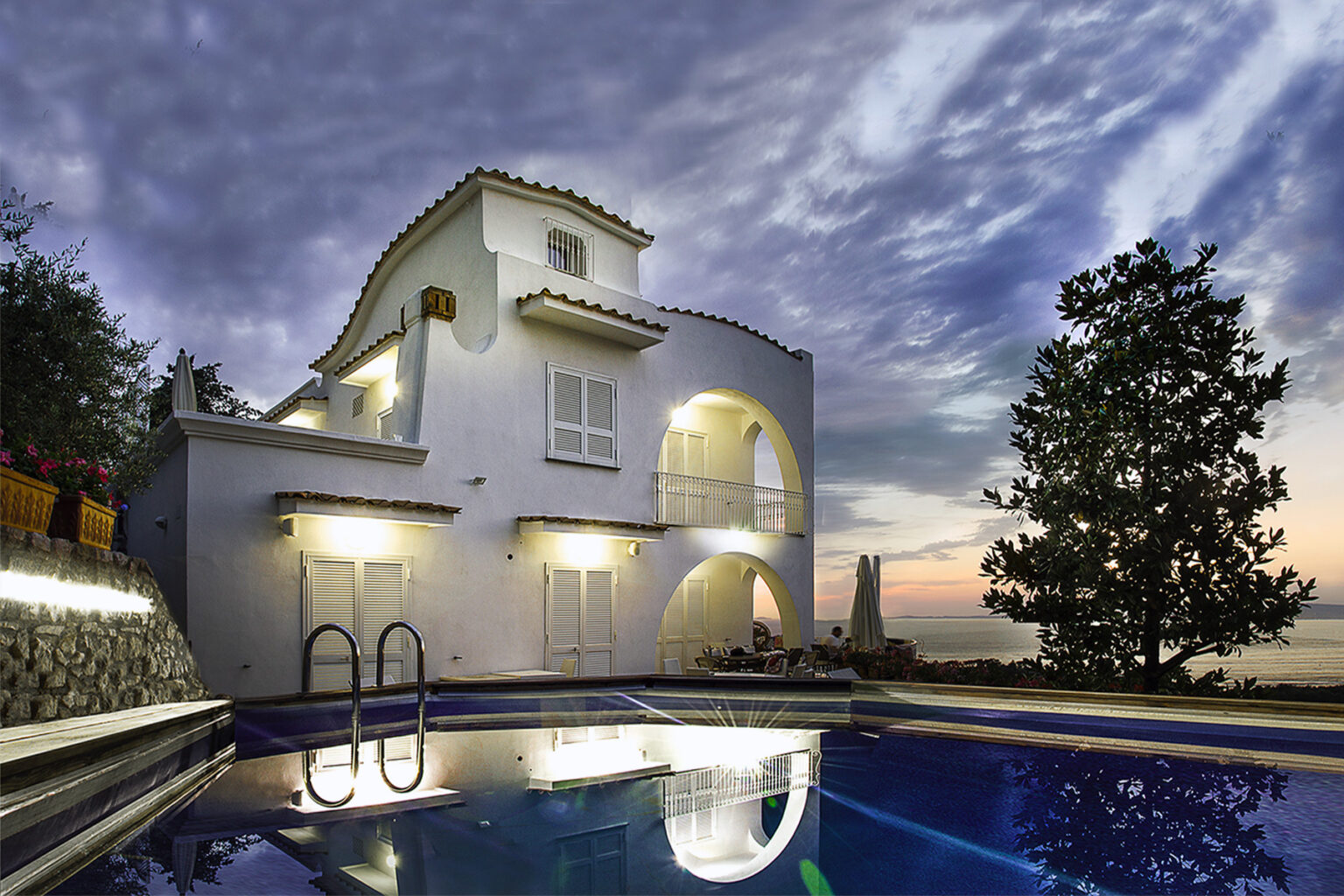 Modern villa with a swimming pool at sunset, featuring a white exterior, large windows, and lush greenery, showcasing a serene coastal atmosphere. | Only on SopranoVillas
