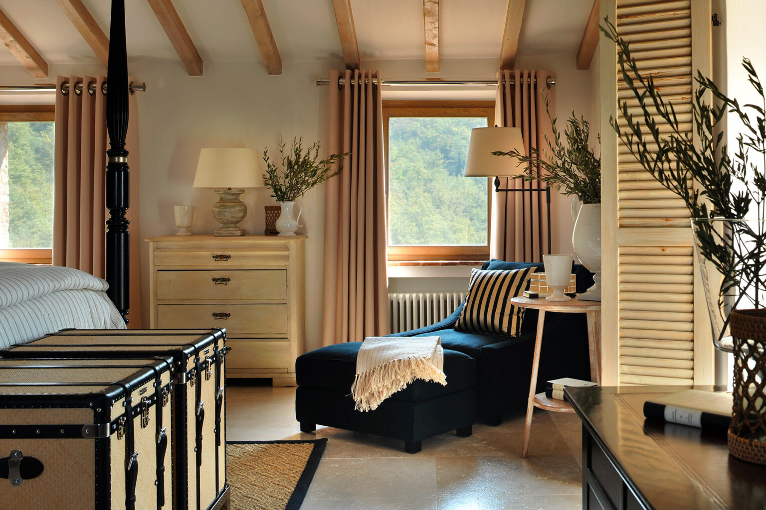 Cozy bedroom interior featuring a black chaise lounge, wooden furniture, and large windows with natural light. Decor includes neutral curtains, a bedside table with a lamp, and decorative plants, creating a warm and inviting atmosphere. | Only on SopranoVillas