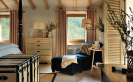 Cozy bedroom interior featuring a black chaise lounge, wooden furniture, and large windows with natural light. Decor includes neutral curtains, a bedside table with a lamp, and decorative plants, creating a warm and inviting atmosphere.