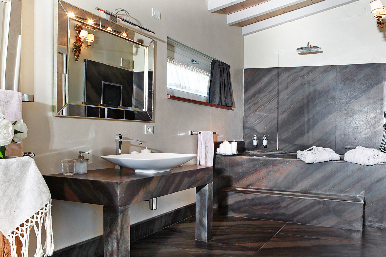 Modern bathroom featuring a sleek stone sink, large mirror, elegant lighting, and a stylish bathtub with plush towels, designed for a luxurious and relaxing atmosphere. | Only on SopranoVillas