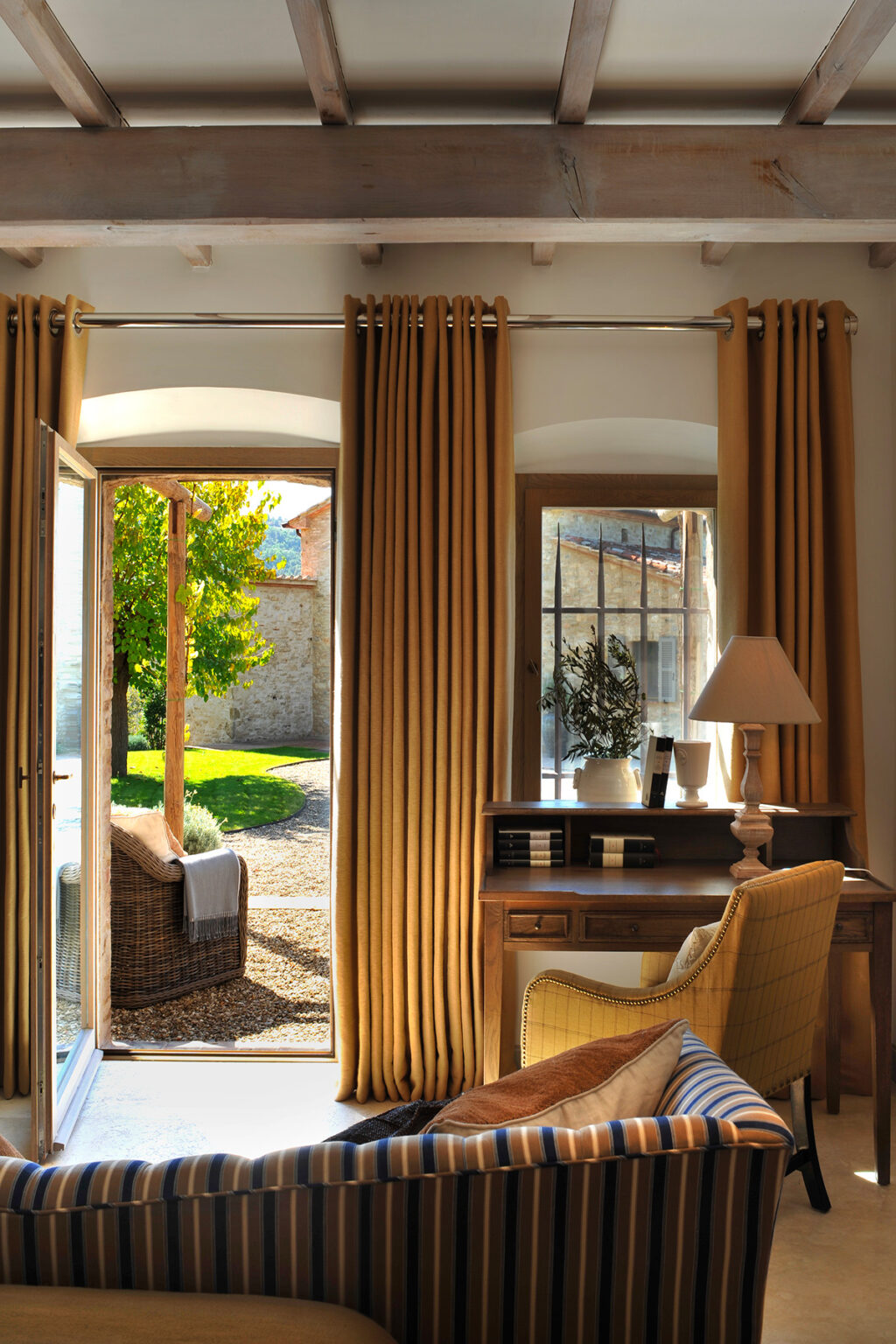 Cozy interior of a sunlit room featuring large windows with golden curtains, a wooden desk, and a lamp, overlooking a lush garden and stone exterior of a rustic building. | Only on SopranoVillas