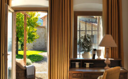 Cozy interior of a sunlit room featuring large windows with golden curtains, a wooden desk, and a lamp, overlooking a lush garden and stone exterior of a rustic building.