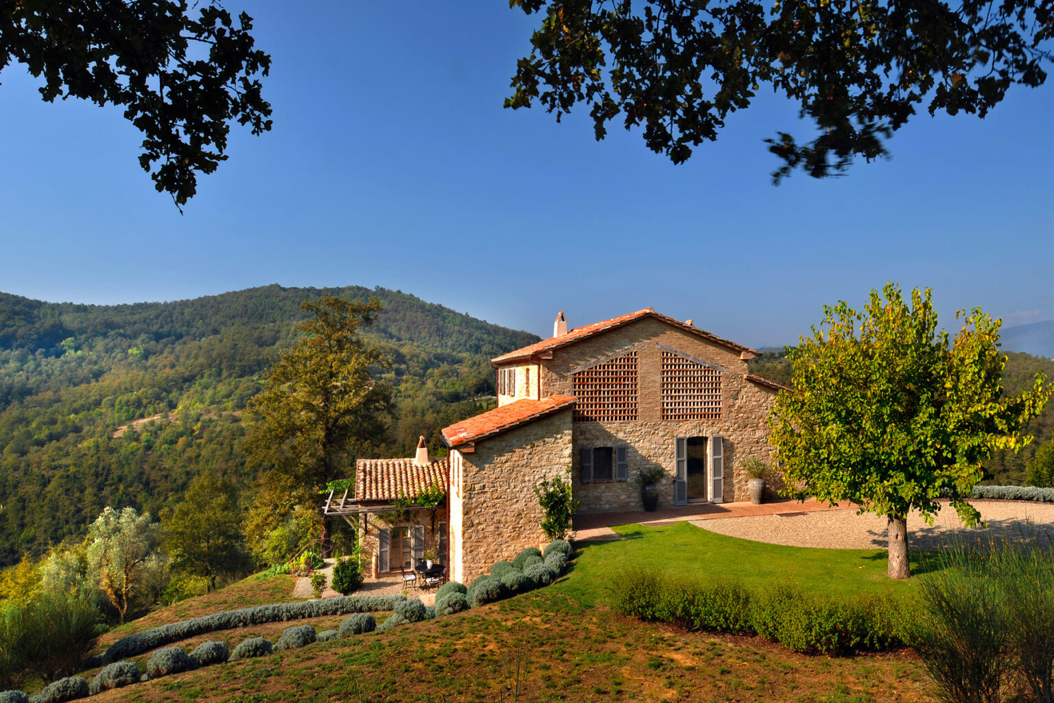 Scenic view of a rustic stone villa nestled in lush green hills, surrounded by trees and vibrant landscaping, showcasing the beauty of countryside architecture in a tranquil setting. | Only on SopranoVillas