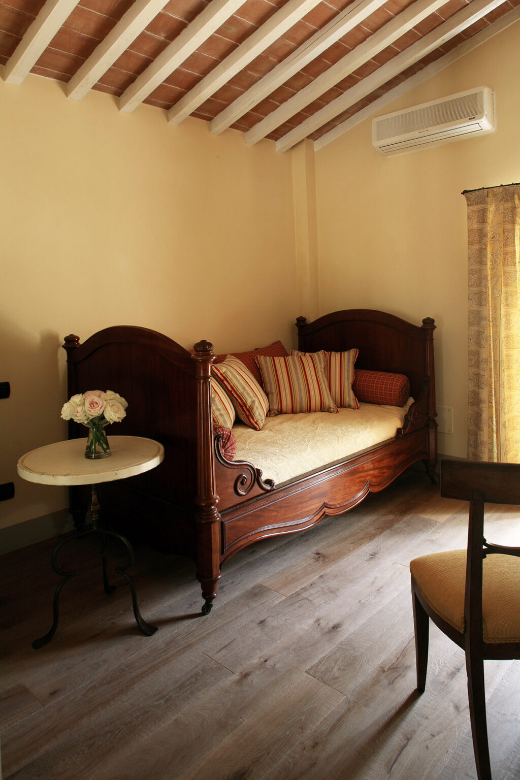 Cozy vintage wooden daybed adorned with decorative cushions, positioned next to a small round table with a vase of fresh flowers, set in a warmly lit room with soft curtains. | Only on SopranoVillas