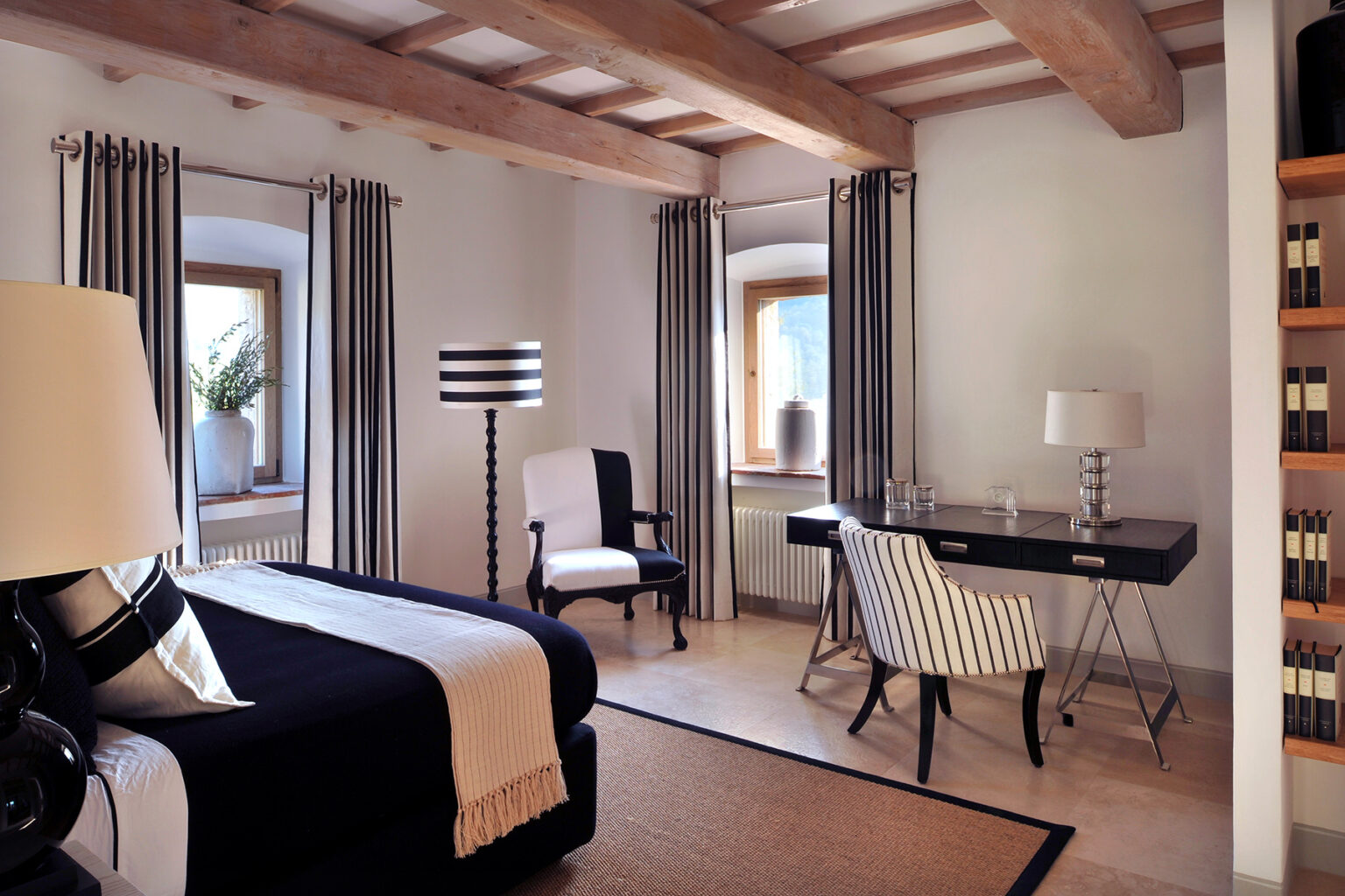Modern bedroom interior featuring wooden beams, stylish furniture, and elegant decor. The room includes a black and white color scheme, a desk with a lamp, and large windows with drapes, creating a bright and inviting atmosphere. | Only on SopranoVillas
