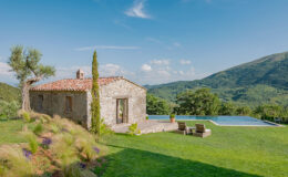 Rustic stone villa with a swimming pool set in a lush green landscape, surrounded by rolling hills and clear blue skies. Perfect for a relaxing getaway in nature.