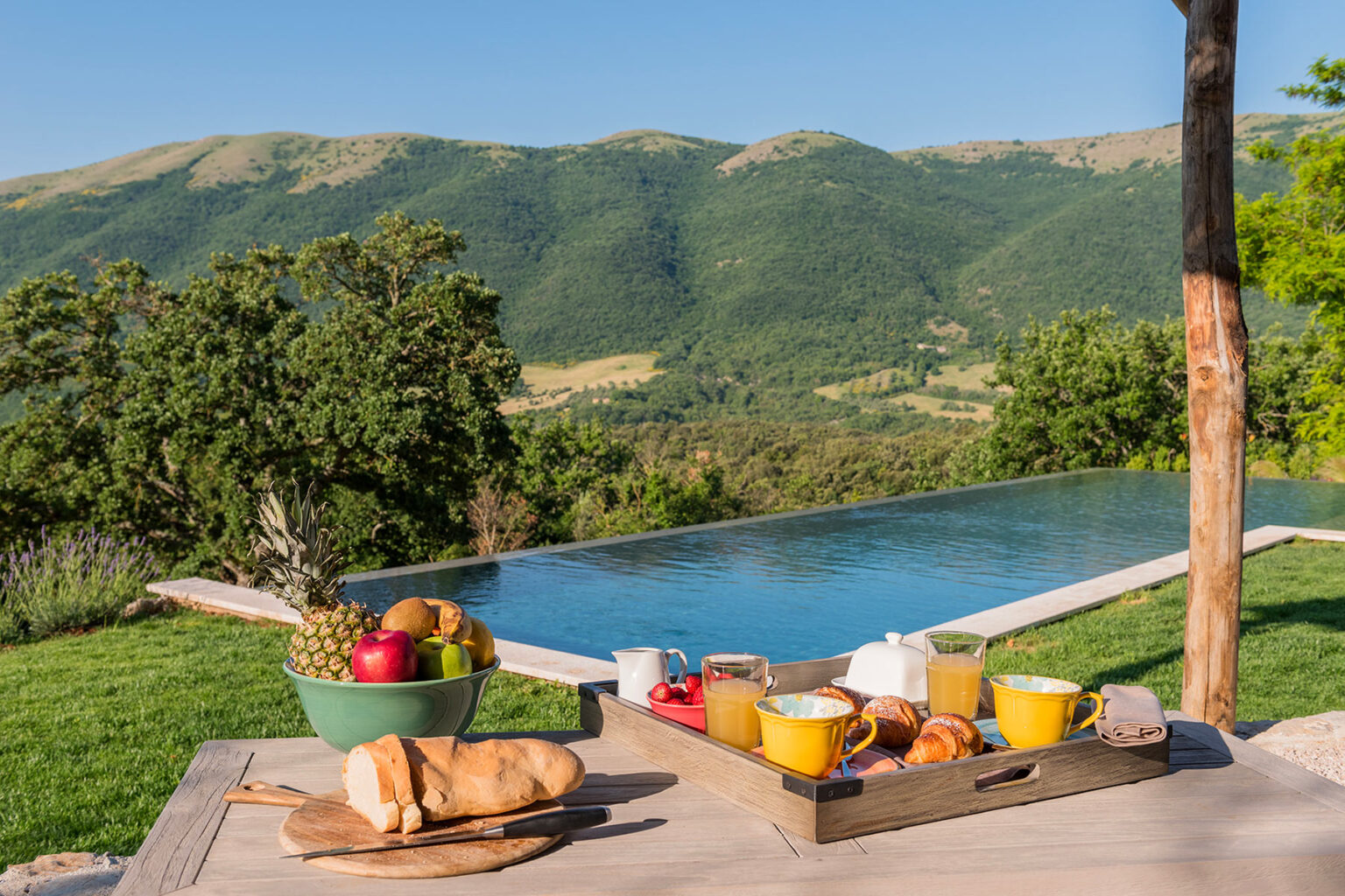 A scenic outdoor breakfast setup featuring a wooden tray with fresh fruits, pastries, and colorful mugs, overlooking a tranquil infinity pool and lush green mountains in the background. | Only on SopranoVillas