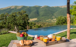 A scenic outdoor breakfast setup featuring a wooden tray with fresh fruits, pastries, and colorful mugs, overlooking a tranquil infinity pool and lush green mountains in the background.