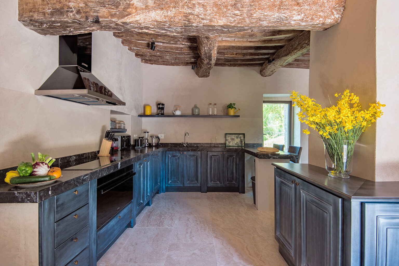 Modern kitchen with rustic wooden beams, dark cabinetry, and a marble countertop, featuring a vase of yellow flowers and fresh fruits on display, creating a warm and inviting atmosphere. | Only on SopranoVillas