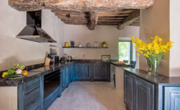 Modern kitchen with rustic wooden beams, dark cabinetry, and a marble countertop, featuring a vase of yellow flowers and fresh fruits on display, creating a warm and inviting atmosphere.