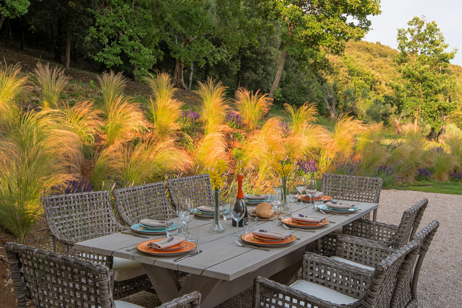 Outdoor dining setup featuring a rustic wooden table surrounded by woven chairs, elegantly arranged with plates, glasses, and a centerpiece of flowers, set against a backdrop of vibrant ornamental grasses and trees. | Only on SopranoVillas