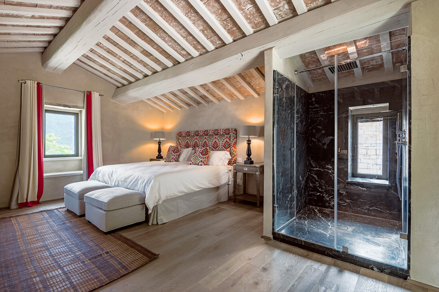 Spacious and stylish bedroom featuring a king-size bed with elegant bedding, two bedside lamps, and a modern glass shower. The room showcases wooden beams, large windows with scenic views, and a cozy area rug, creating a warm and inviting atmosphere. | Only on SopranoVillas