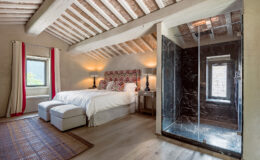 Spacious and stylish bedroom featuring a king-size bed with elegant bedding, two bedside lamps, and a modern glass shower. The room showcases wooden beams, large windows with scenic views, and a cozy area rug, creating a warm and inviting atmosphere.