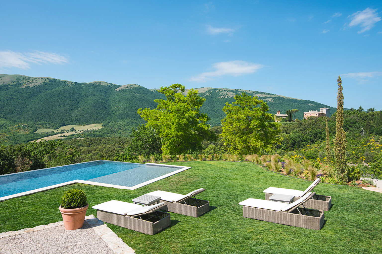 Scenic view of a modern villa with a swimming pool, surrounded by lush green lawns and trees, set against a backdrop of rolling hills and blue skies. Loungers are arranged by the pool, creating an inviting outdoor relaxation space. | Only on SopranoVillas