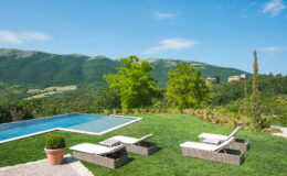 Scenic view of a modern villa with a swimming pool, surrounded by lush green lawns and trees, set against a backdrop of rolling hills and blue skies. Loungers are arranged by the pool, creating an inviting outdoor relaxation space.
