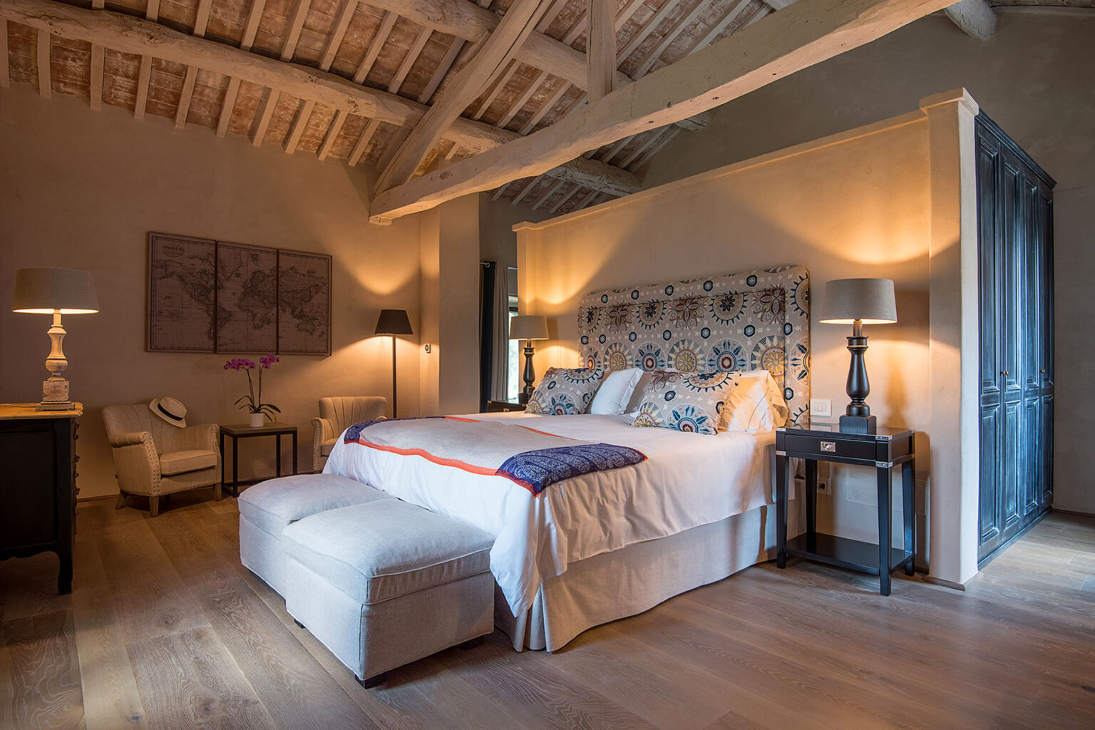Spacious and elegantly designed bedroom featuring a king-size bed with decorative pillows, a stylish upholstered headboard, and warm lighting. The room includes a cozy seating area with an armchair, a bedside table, and wooden flooring, creating a serene and inviting atmosphere. | Only on SopranoVillas