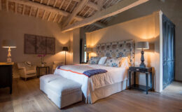 Spacious and elegantly designed bedroom featuring a king-size bed with decorative pillows, a stylish upholstered headboard, and warm lighting. The room includes a cozy seating area with an armchair, a bedside table, and wooden flooring, creating a serene and inviting atmosphere.