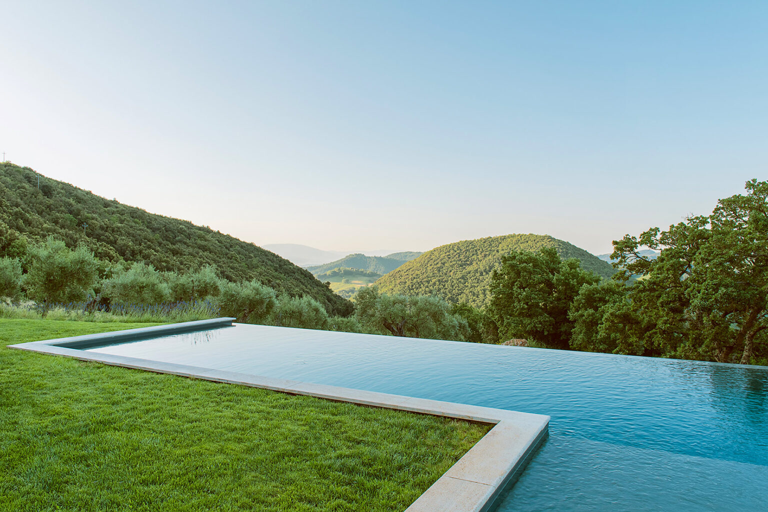 Infinity pool overlooking lush green hills under a clear blue sky, surrounded by olive trees, creating a serene and tranquil outdoor setting. | Only on SopranoVillas