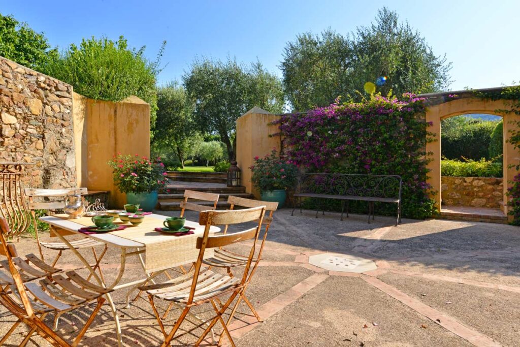 Charming outdoor garden area featuring a rustic stone wall, potted plants, and a cozy table set with chairs, creating a serene atmosphere for relaxation and dining. Bright sunlight enhances the vibrant greenery and colorful flowers, ideal for a peaceful retreat. | Only on SopranoVillas