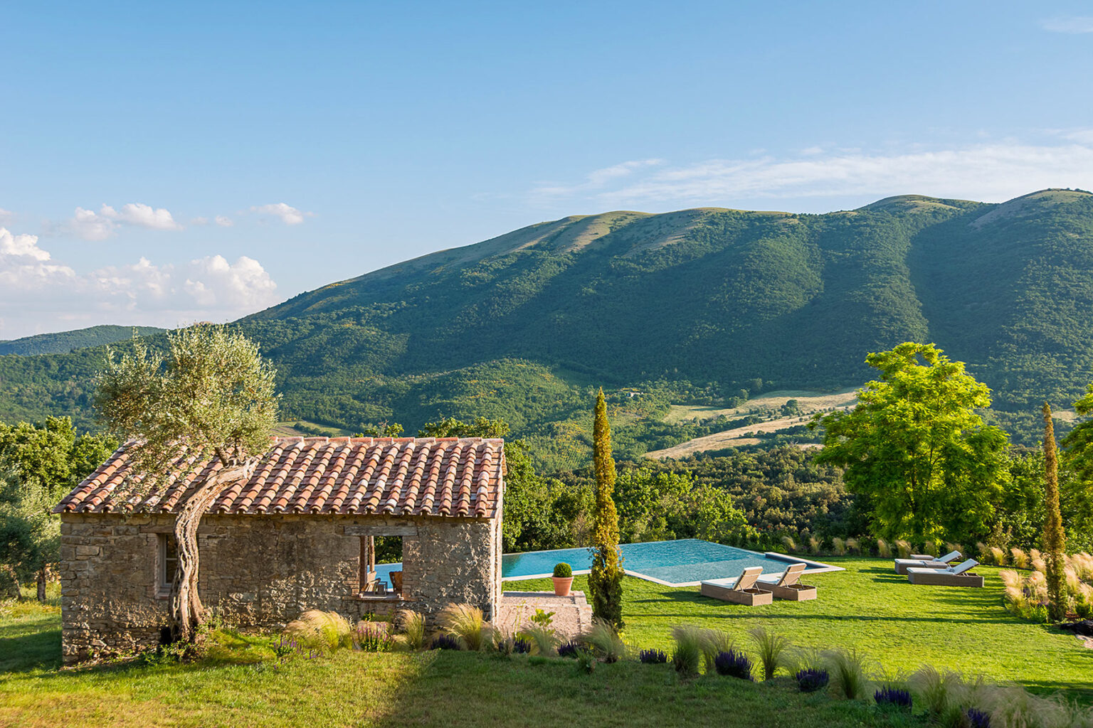 Scenic view of a rustic stone cottage surrounded by lush greenery, with a swimming pool in the foreground and rolling hills in the background under a clear blue sky. Perfect for vacation rentals or nature retreats. | Only on SopranoVillas