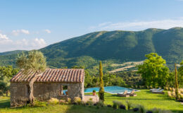 Scenic view of a rustic stone cottage surrounded by lush greenery, with a swimming pool in the foreground and rolling hills in the background under a clear blue sky. Perfect for vacation rentals or nature retreats.