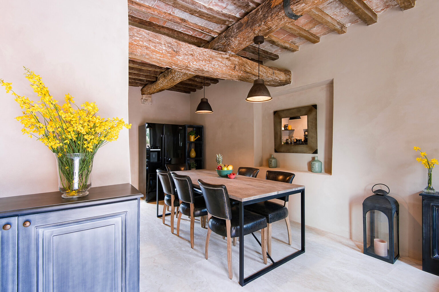 Modern dining room featuring a wooden table with black chairs, rustic exposed wooden beams, and a stylish mirror. A vase of yellow flowers adds a touch of color, complemented by minimalist decor and a black refrigerator. | Only on SopranoVillas