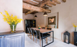 Modern dining room featuring a wooden table with black chairs, rustic exposed wooden beams, and a stylish mirror. A vase of yellow flowers adds a touch of color, complemented by minimalist decor and a black refrigerator.