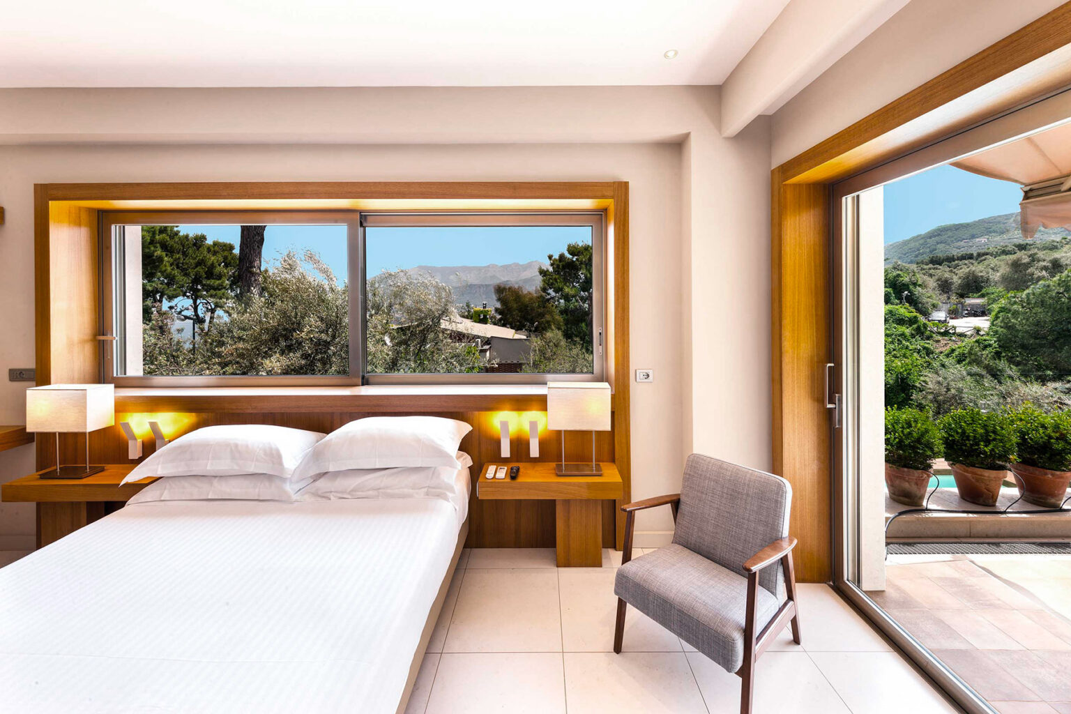 Modern hotel room featuring a comfortable bed, stylish wooden furniture, and a large window with scenic views. The room includes bedside lamps and a cozy chair, with an open door leading to a balcony and outdoor greenery. Perfect for relaxation and enjoying nature. | Only on SopranoVillas