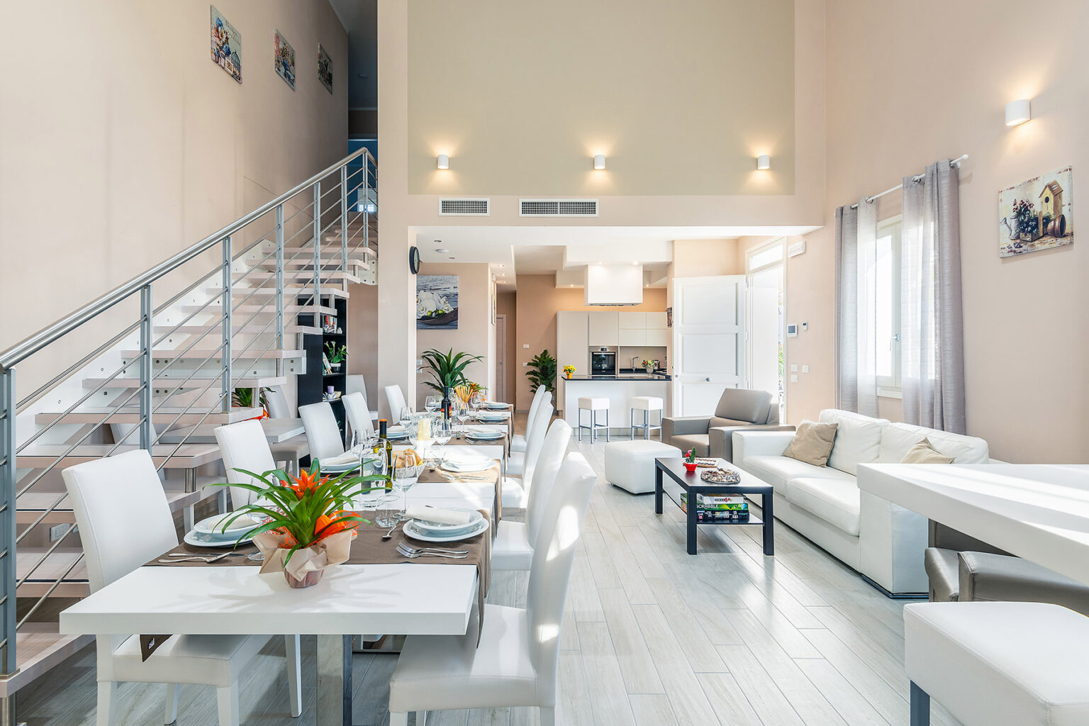Modern open-concept living space featuring a stylish dining area with a long table set for dinner, a cozy lounge with a white sofa, and a sleek kitchen visible in the background. The high ceilings and large windows create a bright and airy atmosphere. | Only on SopranoVillas