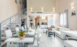 Modern open-concept living space featuring a stylish dining area with a long table set for dinner, a cozy lounge with a white sofa, and a sleek kitchen visible in the background. The high ceilings and large windows create a bright and airy atmosphere.