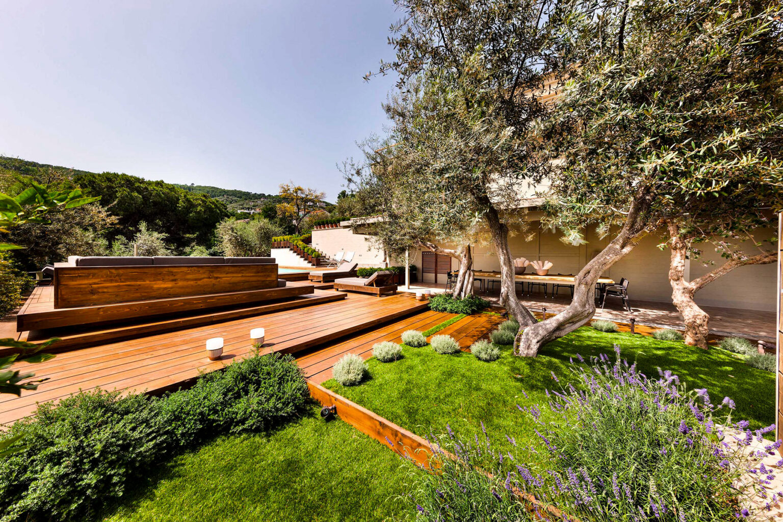 Modern outdoor garden featuring wooden decking, lush green grass, and olive trees, ideal for relaxation and entertaining in a serene natural setting. | Only on SopranoVillas