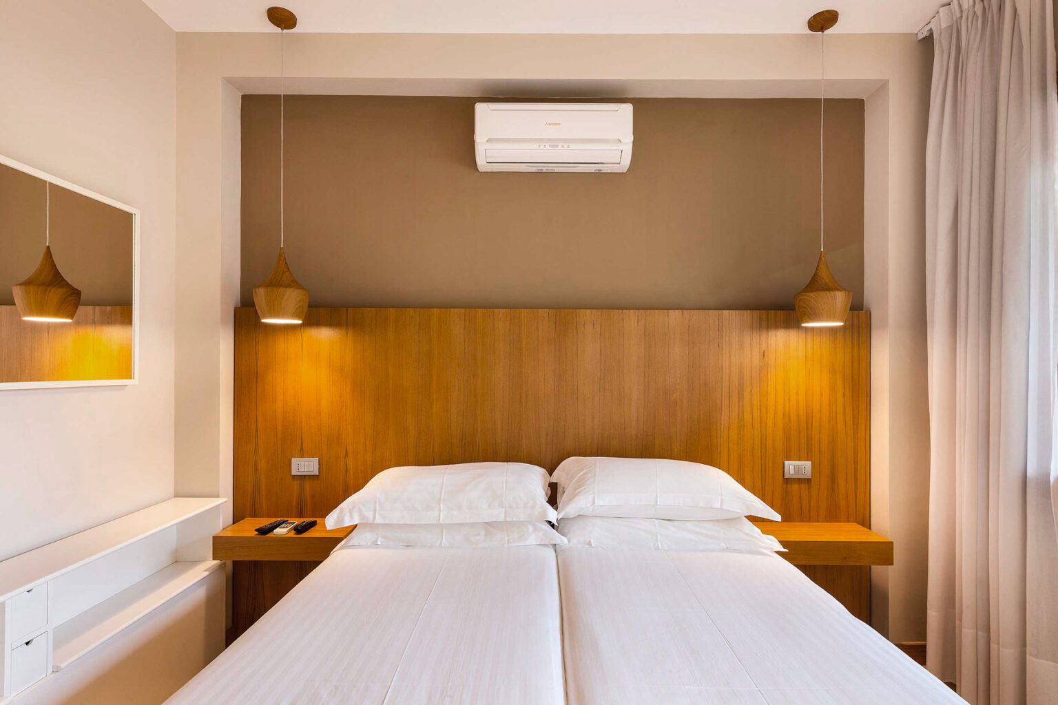 Modern hotel room featuring a stylish twin bed setup with a wooden headboard, pendant lighting, an air conditioning unit, and a minimalist design aesthetic. | Only on SopranoVillas
