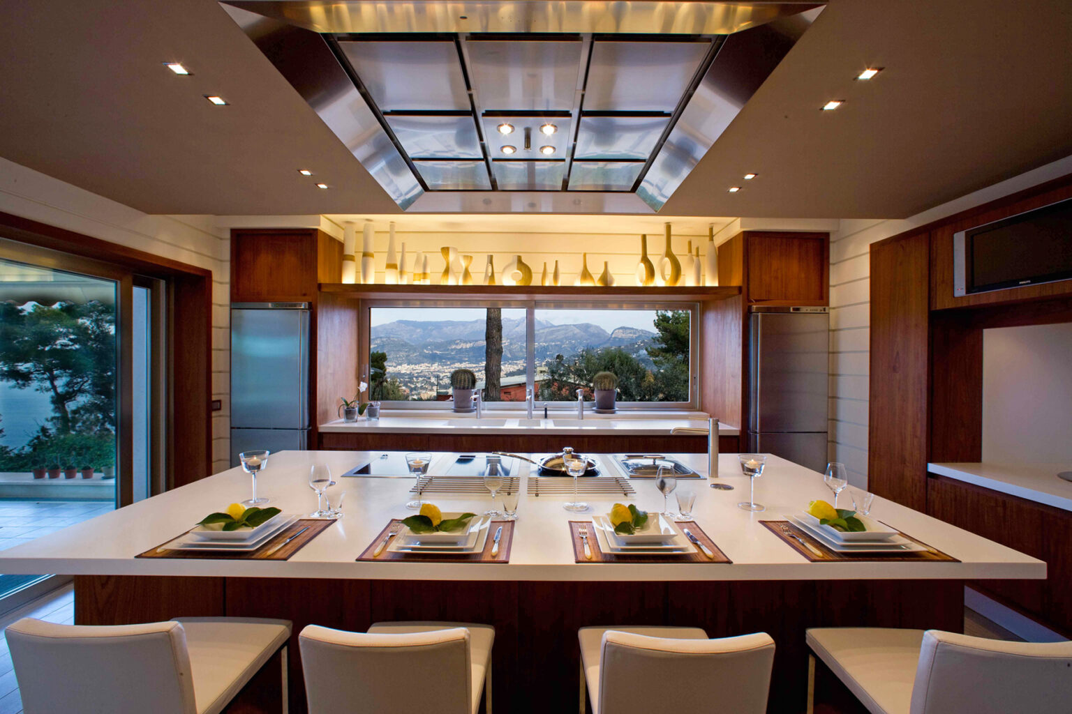 Modern kitchen interior featuring an elegant island with seating, contemporary cabinetry, and large windows showcasing a scenic view. The design incorporates warm wood tones and sophisticated lighting, creating a stylish and inviting atmosphere. | Only on SopranoVillas