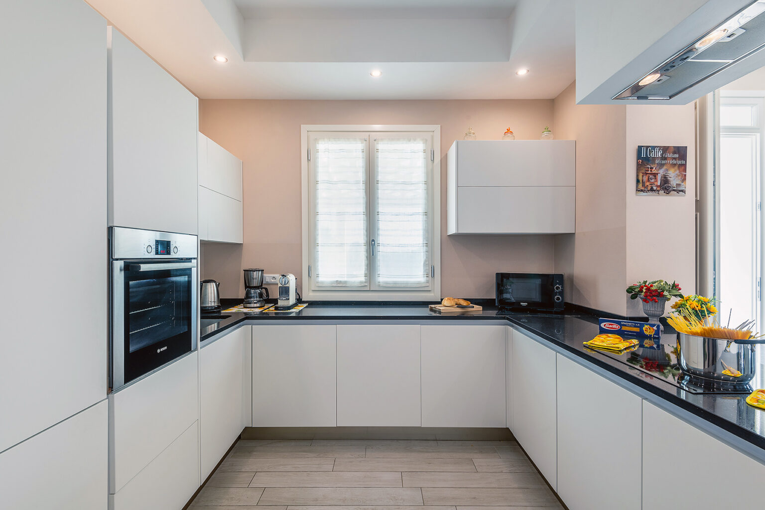 Modern kitchen design featuring sleek white cabinetry, a dark countertop, built-in oven, and ample natural light from a window. Stylish appliances and decorative elements enhance the contemporary aesthetic. | Only on SopranoVillas