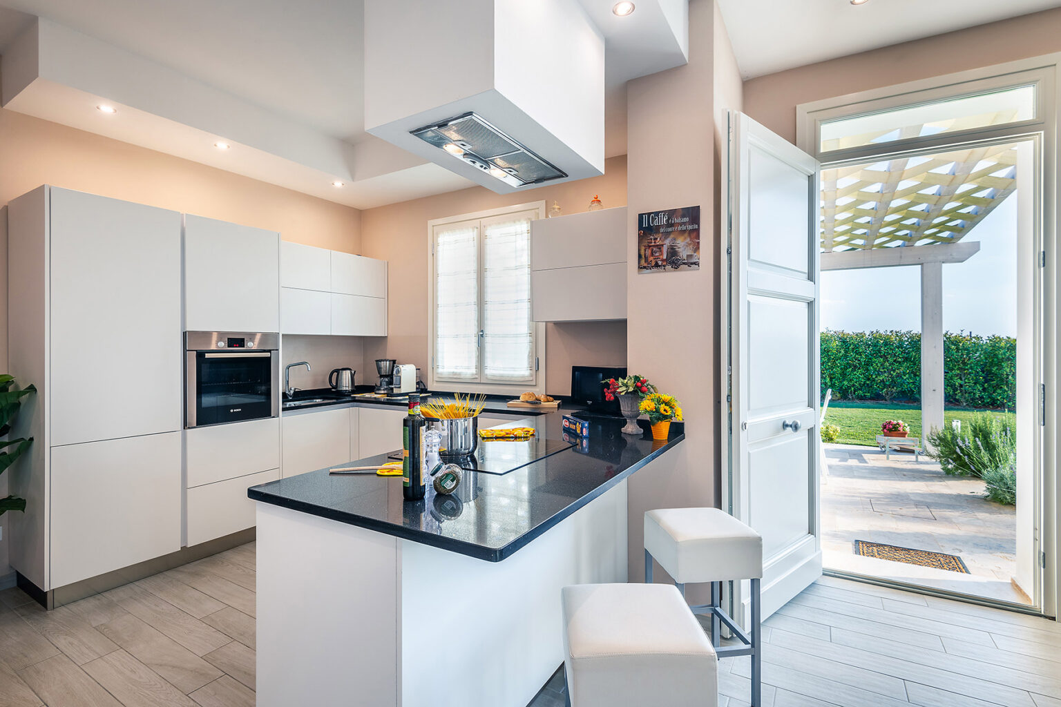 Modern kitchen interior featuring sleek white cabinetry, a black countertop, and stainless steel appliances. The space is well-lit with natural light streaming in from an open door leading to a garden, creating a bright and inviting atmosphere. | Only on SopranoVillas