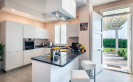Modern kitchen interior featuring sleek white cabinetry, a black countertop, and stainless steel appliances. The space is well-lit with natural light streaming in from an open door leading to a garden, creating a bright and inviting atmosphere.