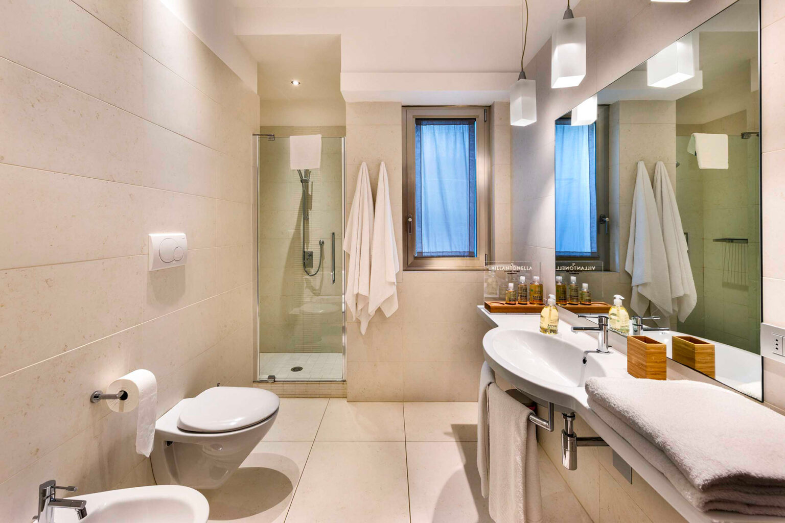 Modern bathroom with neutral tones, featuring a shower area, sleek toilet, and a double sink vanity with wooden accents. Natural light filters through a window, enhancing the spacious feel of the room. | Only on SopranoVillas