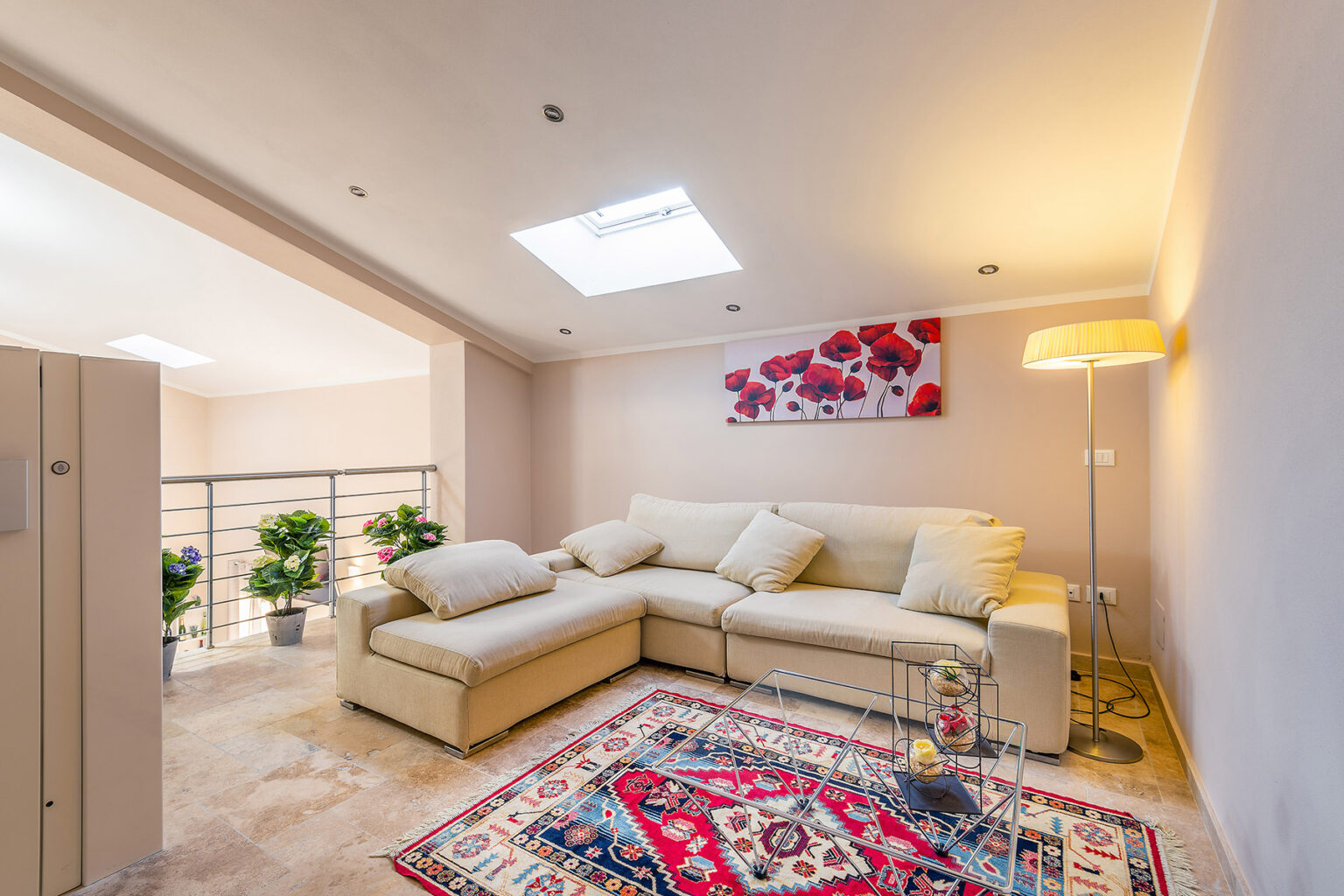 Cozy living room featuring a light-colored sectional sofa, a decorative rug, and modern lighting. The space is enhanced by natural light from a skylight and includes potted plants and wall art. Ideal for relaxation and contemporary interior design. | Only on SopranoVillas