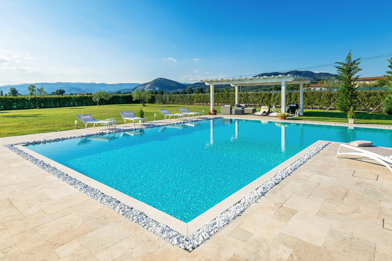 Modern outdoor swimming pool surrounded by a spacious patio, lounge chairs, and lush green landscape with mountains in the background. Ideal for relaxation and leisure activities. | Only on SopranoVillas