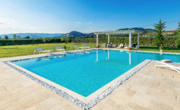 Modern outdoor swimming pool surrounded by a spacious patio, lounge chairs, and lush green landscape with mountains in the background. Ideal for relaxation and leisure activities.